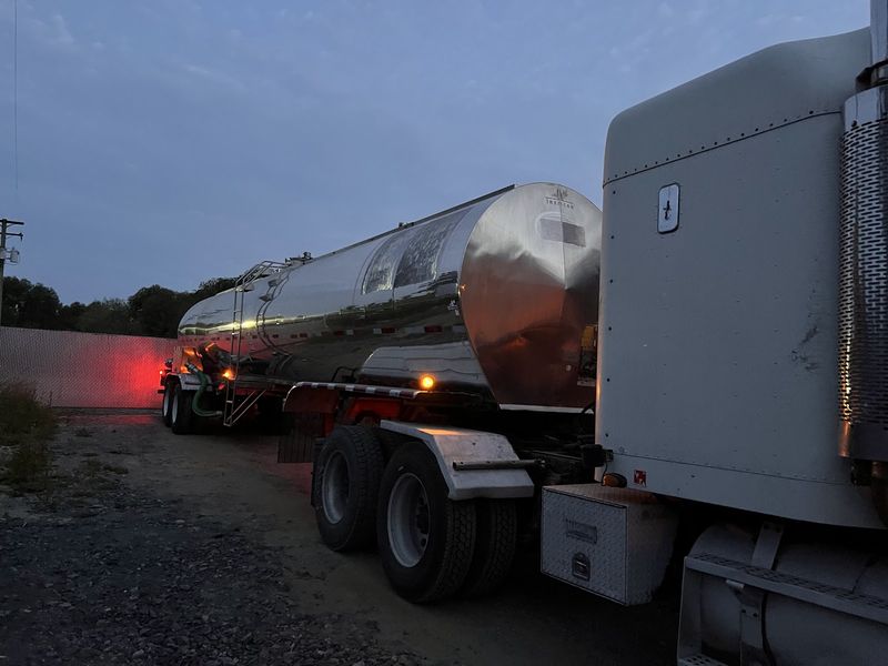 Tractor and tanker trailer in a gravel yard at dawn, tail lights glowing red.