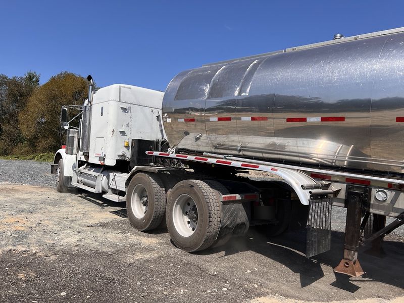 White tractor hitched to the polished tanker trailer on gravel under a blue sky.